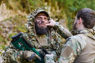 Obraz premium Bearded Soldier In Camouflage Receiving Face Paint Before Field Operation In Forest Training