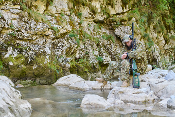 Obraz premium Sniper In Camouflage Sitting On Rocky Riverbank With Rifle In Woodland Ambush