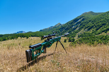 Fototapeta premium Bolt-Action Sniper Rifle With Scope And Bipod On Dry Grass Overlooking Mountain Valley