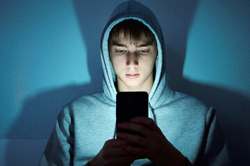 Teenage boy in a hoodie sitting in the dark, illuminated by the glow of his smartphone while looking at the screen © Pihuliak