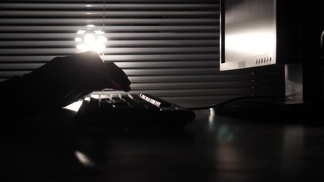 Person's hands typing on a computer keyboard in a dark room, creating a silhouette effect against bright light and window blinds, suggesting late night work or cyber activity
