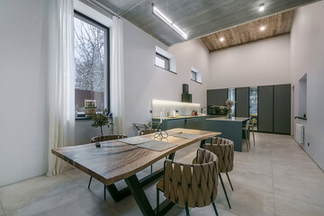 Fototapeta premium A dining table with chairs sits in a modern kitchen. Large windows let in light and show a view of trees outside. The space is open and clean with simple design features