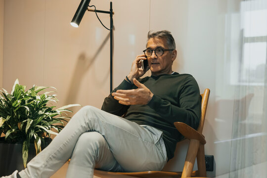 Full length of senior male entrepreneur talking on smart phone while sitting on chair at office seen through glass