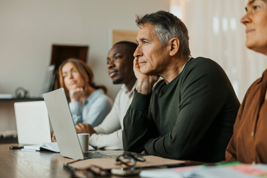 Senior businessman leaning on elbow while sitting in meeting with colleagues at office