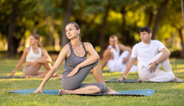 Calm fit woman performing twisting asana Matsyendrasana, or Lord of Fishes Pose, sitting on mat on green grassy glade in summer park during daily group yoga session