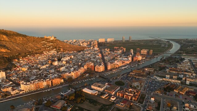 Aerial drone view of Cullera in Spain during golden hour, featuring a hilltop castle and winding path above the Mediterranean Sea. Detailed coastal cityscape with rooftops, shoreline and dramatic suns