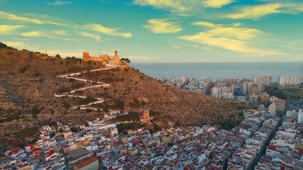 Obraz premium Aerial drone view of Cullera in Spain during golden hour, featuring a hilltop castle and winding path above the Mediterranean Sea. Detailed coastal cityscape with rooftops, shoreline and dramatic suns