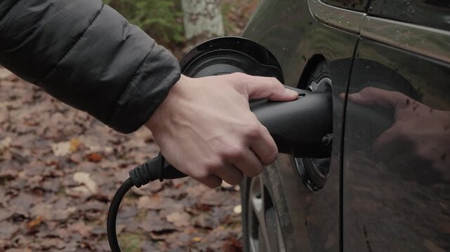 Electric vehicle disconnection. Hand unplugs charger from vehicle. Charging electric vehicle beside muddy leafcovered roadside. Person disconnecting charger from car on rural path