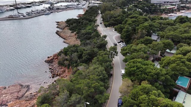 Aerial view of coastal road with cars near Temple of Apollo Zoster at Vouliagmeni peninsula with yacht marina and lush greenery, Mediterranean Greece