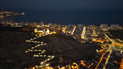 Obraz premium Aerial night view of Cullera in Spain featuring illuminated hilltop castle and glowing city streets along the Mediterranean coast. Detailed coastal skyline with fortress walls and seaside urban landsc