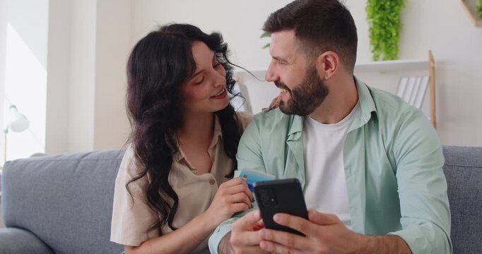 Couple making online payment with credit card and mobile phone app. Young happy woman and man sitting on home sofa to use smartphone to confirm banking details and complete secure shopping checkout