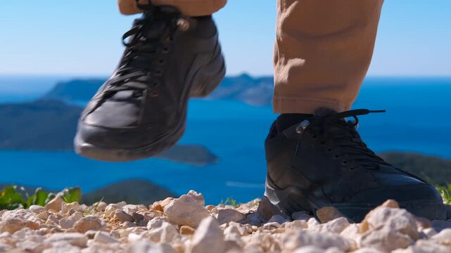 Hiker stomping feet on a mountain top overlooking the sea. Close-up of a hiker's feet in black boots stomping on a rocky mountain path, celebrating reaching the summit with a view of the blue sea