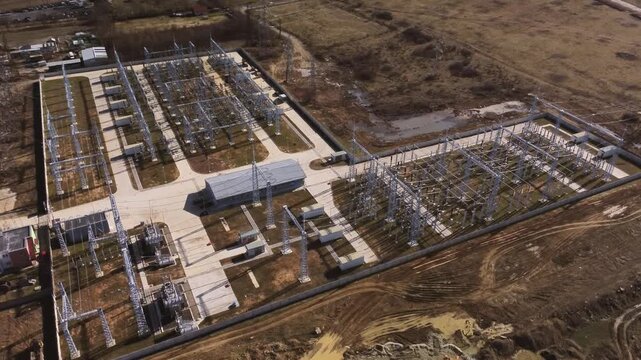 Aerial drone view of high-voltage electrical substation with transformers and switchgear, power lines and pylons, fenced industrial grid facility in rural landscape.