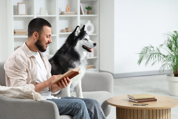 Handsome young man sitting on sofa and reading book with cute husky dog at home © Pixel-Shot
