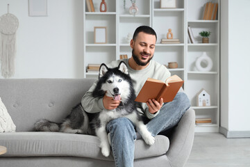 Handsome young man sitting on sofa and reading book with cute husky dog at home © Pixel-Shot