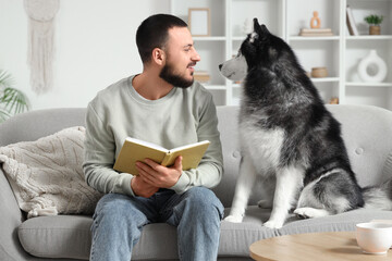 Handsome young man sitting on sofa and reading book with cute husky dog at home © Pixel-Shot