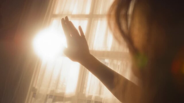 Cinematic POV shot of young woman waving hand against bright sunlight shining through curtains creating soft warm lighting with lens flare, copy space