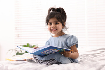 Cute little girl reading book on bed at home