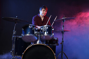 Smiling teenage boy with drumsticks learning how to play drums in color lights and smoke against dark background