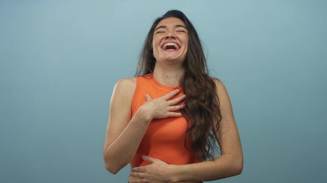 Woman laughs heartily with hand on chest and long brown hair falling over shoulders, wearing an orange sleeveless top against a light blue studio wall; joy.