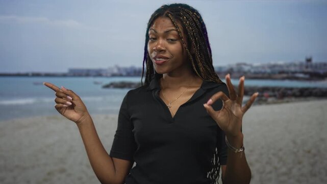 Woman points finger and makes ok sign with hand on a sandy beach near a rocky jetty and shoreline, smiling with braided hair and visible necklace; joyful confidence.