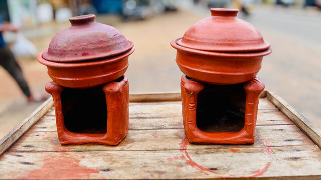 Two traditional handmade terracotta clay stoves with lidded cooking pots resting on a rustic wooden board with a blurred outdoor background.
