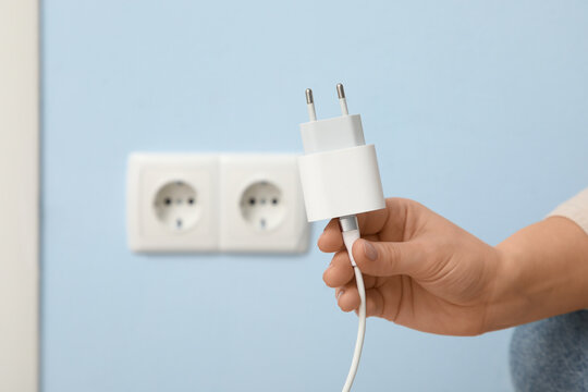 Female hand with unplugged charger near socket on blue wall at home, closeup