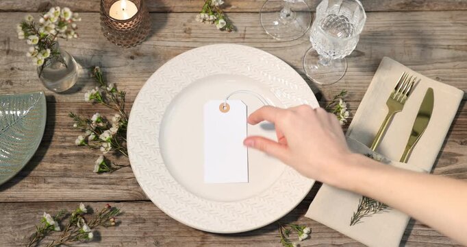 Stylish table setting. Woman putting card, floral decor and burning candle on wooden background, above view