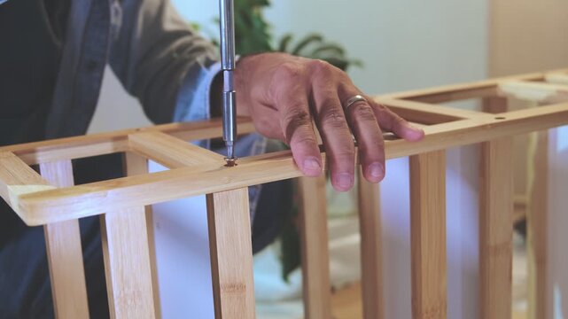 Unrecognizable man assembling a new piece of wooden furniture on the floor with a screwdriver