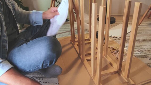 Young man sitting on the floor assembling new wooden furniture and following the instructions