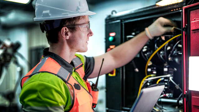 A male engineer in a hard hat holds a laptop while inspecting electrical wires. He is a specialist doing maintenance on a machine in a high-tech smart factory.