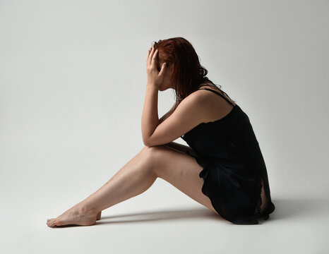 full length portrait of beautiful female model, wet messy hair, simple black silk robe gown, bruised legs. sitting pose with emotive sad pose. isolated figure studio background, dark contrast shadows