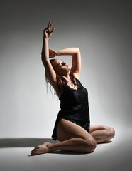 Obraz premium full length portrait of beautiful girl model, wet messy hair, simple black silk robe gown, bruised leg. sitting pose with emotive hand gesture reaching. isolated figure studio background dark shadows