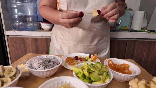 A woman's hands are preparing a fruit kebab, stringing pieces of fruit on skewers