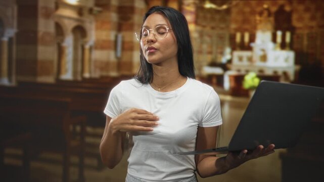 Woman holding laptop and touching chest with right hand inside church while gesturing and speaking; contemplation faith.