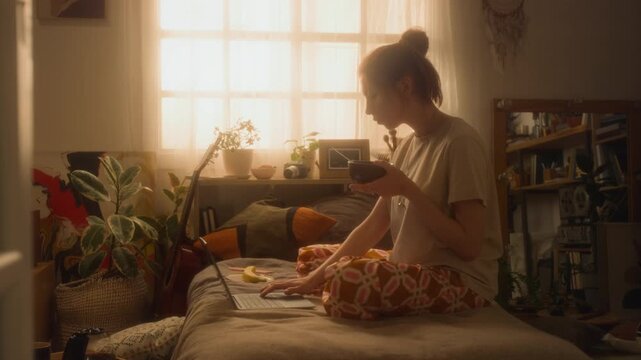 Cinematic side view shot of young woman using laptop on bed and eating breakfast while enjoying slow morning routine at home backlit by warm sunlight, copy space