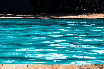 Crystal clear turquoise water ripples gently in a sunlit outdoor swimming pool, reflecting bright sunlight on its surface with a stone tiled edge visible in the foreground and background