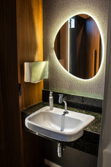 Modern bathroom interior featuring a white ceramic washbasin on a dark granite countertop, an organic-shaped backlit mirror on a textured wall, and warm wooden paneling in a luxury hotel setting