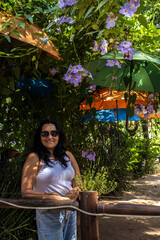 A smiling woman relaxes in a beautiful garden cafe, creatively decorated with colorful hanging umbrellas and lush flowering vines on a sunny day. 