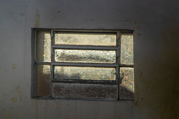 Sunlight glows through an old textured glass window in a dark, cracked wall. A silhouette reflects on the vintage pane. Rustic Brazilian architecture showcasing light, shadow, and urban isolation. 