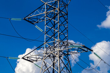 High-voltage electricity transmission tower with glass insulators against a vibrant blue sky with white clouds. Symbol of the power grid, energy distribution, and industrial infrastructure.