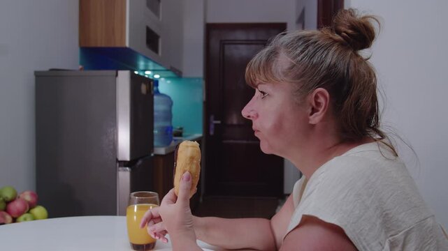 A woman eats a fresh donut covered with icing and washes it down with juice, side view