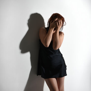 Close portrait of girl model, wet messy hair wearing simple black silk robe gown, standing pose leaning against a wall with strained emotive pose. Isolated figure with shadows on studio background.