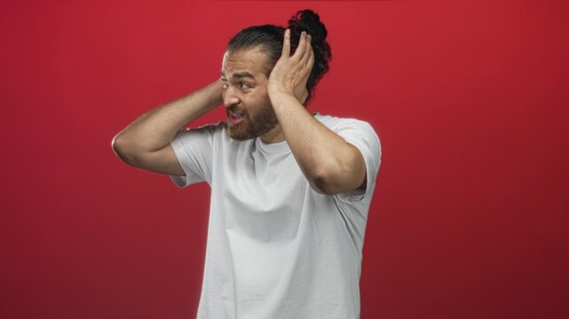 Man in white t shirt pressing hands to ears and grimacing in a red studio, covering ears with both hands and showing beard and clenched jaw; distress.