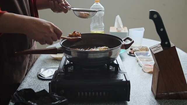 Close-up of home chef hands draining golden corn fritters using wooden spoon and strainer in pan on portable stove
