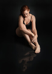 Naklejka premium full length female model with damp hair, wearing simple black glamour dress. sitting kneeling pose on wet black floor with water reflections. isolated figure on dark studio background.