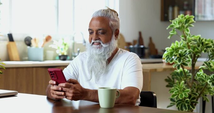 Indian mid age stylish man with long white beard and hair using smartphone while sitting at breakfast table in modern kitchen interior, browsing internet, chatting online at home