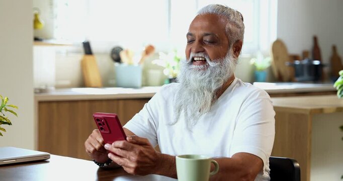 Indian mid age stylish man with long white beard and hair using smartphone while sitting at breakfast table in modern kitchen interior, browsing internet, chatting online at home