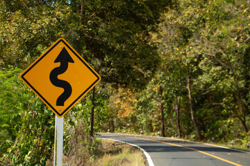 Yellow winding road warning sign beside a curving asphalt road through a lush forest landscape in natural daylight.