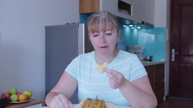 A woman sits at a table in front of a kitchen and eats a pineapple